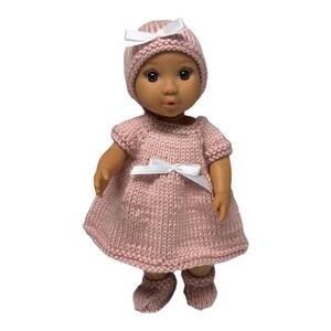 May include: A doll with dark skin and brown eyes wearing a hand-knitted light pink dress, a matching bonnet, and booties. The dress and bonnet are accented with small white satin ribbons. The knitted fabric has a consistent texture throughout.
