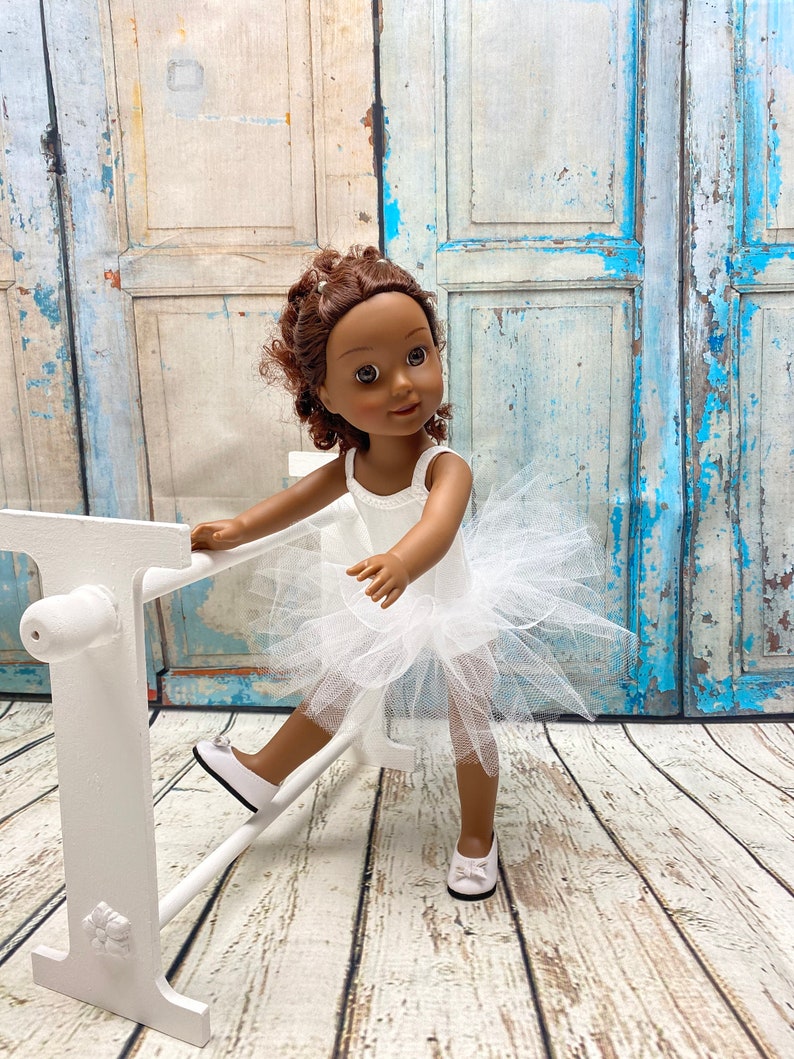 Pu&ograve; includere: Una bambola dalla pelle scura che indossa un tut&ugrave; bianco e scarpette da ballet bianche, tenendosi a una sbarra da ballet in legno bianco.