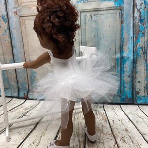 Pu&ograve; includere: Una bambola con un tut&ugrave; di tulle bianco e un body bianco. La bambola ha i capelli ricci castani e sta in piedi davanti a una superficie di legno.