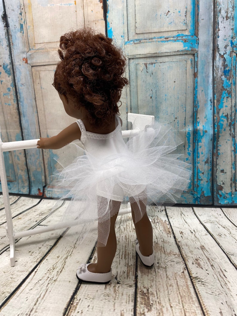 Pu&ograve; includere: Una bambola con un tut&ugrave; di tulle bianco e un body bianco. La bambola ha i capelli ricci castani e sta in piedi davanti a una superficie di legno.