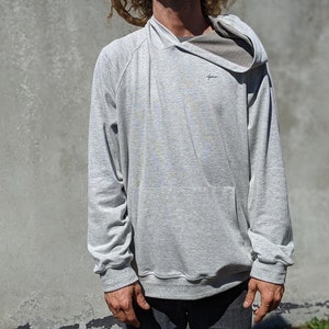 Puede incluir: Una sudadera gris con capucha y un pequeño logotipo en la parte delantera. La sudadera tiene un corte holgado y mangas largas.