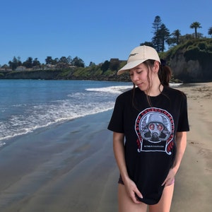 Puede incluir: Una persona lleva una camiseta negra con un gráfico de una persona con una máscara de gas y el texto "Stop Geoengineering" y el sitio web "actforlactivism.com". La persona lleva una gorra de béisbol beige con la palabra "Pro" en ella.