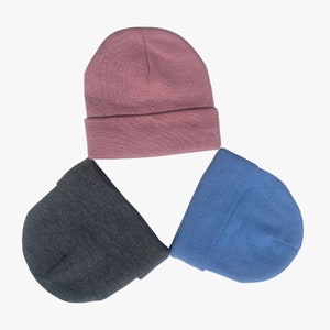 Puede incluir: Tres gorros de punto en disposición triangular. Los gorros son en tonos rosa empolvado, gris antracita y azul claro. Cada gorro tiene un dobladillo doblado en la base. Los sombreros están hechos de un material de punto suave.