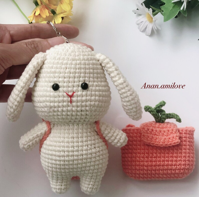 May include: White crocheted bunny with a red nose and black eyes, carrying a small orange crocheted carrot bag. Anan.amilove is written in the background.