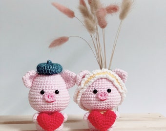 May include: Two crocheted pink pig figurines holding red hearts. The pig on the left is wearing a blue beret and the pig on the right is wearing a yellow and white hat. The pigs are sitting on a wooden surface.