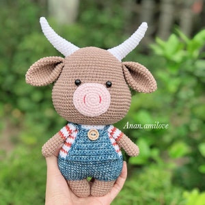 Op de afbeelding: Een bruin gehaakt knuffeldier in de vorm van een koe met witte hoorns. De koe draagt een blauwe denim overall met rode en witte strepen op de bovenkant. De koe heeft een roze neus en zwarte ogen.