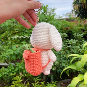 May include: White crocheted bunny keychain with a small orange crocheted backpack. The backpack has a green leafy stem sticking out of the top.