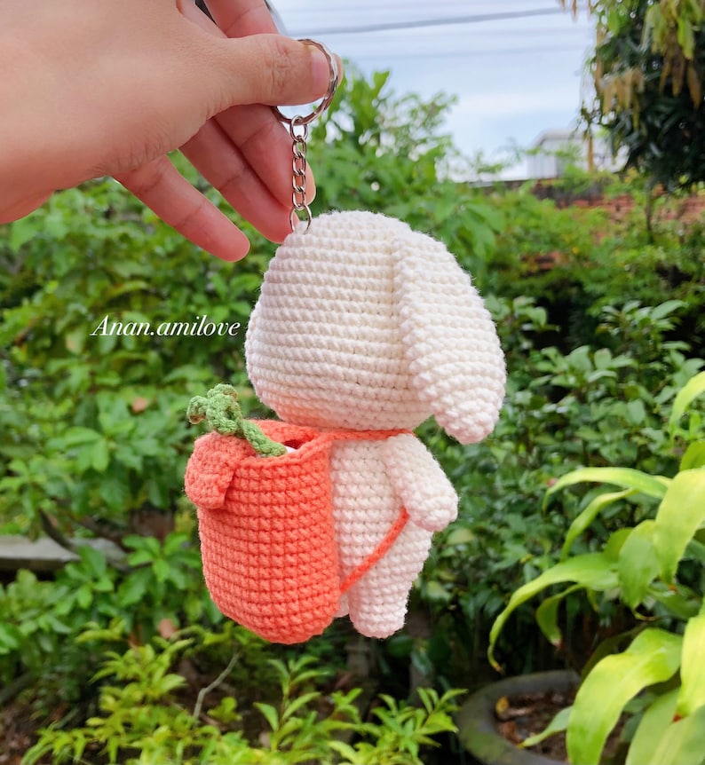 May include: White crocheted bunny keychain with a small orange crocheted backpack. The backpack has a green leafy stem sticking out of the top.