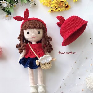 Peut inclure: Une poup&eacute;e en crochet aux cheveux bruns, portant une robe rouge, une jupe bleue et un sac &agrave; main en forme d'agneau blanc en crochet. La poup&eacute;e tient un chapeau rouge en crochet avec des oreilles de lapin. La poup&eacute;e est assise sur une surface blanche avec un fond blanc.