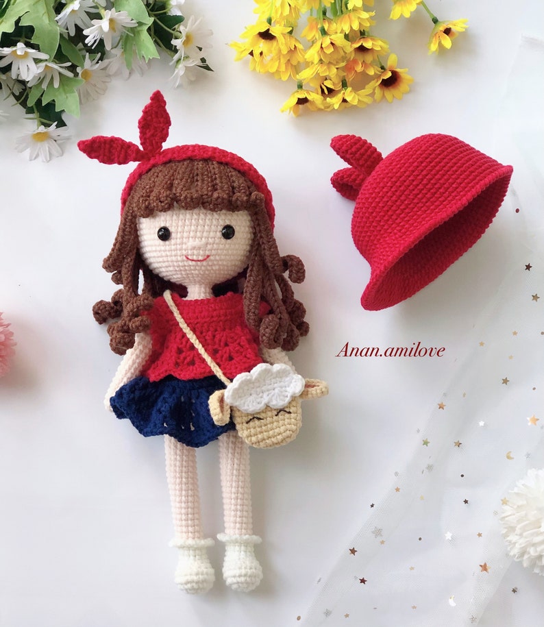 Peut inclure: Une poup&eacute;e en crochet aux cheveux bruns, portant une robe rouge, une jupe bleue et un sac &agrave; main en forme d'agneau blanc en crochet. La poup&eacute;e tient un chapeau rouge en crochet avec des oreilles de lapin. La poup&eacute;e est assise sur une surface blanche avec un fond blanc.
