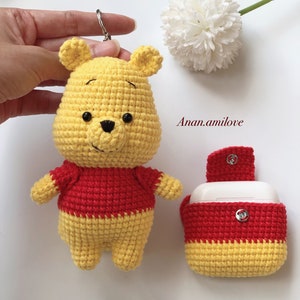 May include: A crocheted Winnie the Pooh bear keyring with a red shirt and a matching crocheted case for wireless earbuds. The case has a small silver button.