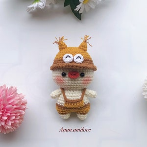 May include: A crocheted toy with a white body, brown overalls, and a brown hat with white eyes. The toy has a red nose and pink cheeks.