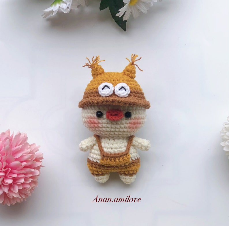 May include: A crocheted toy with a white body, brown overalls, and a brown hat with white eyes. The toy has a red nose and pink cheeks.