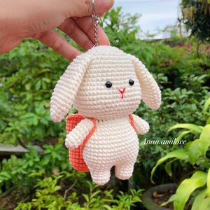 May include: White crocheted bunny keychain with a small orange backpack. The bunny has black eyes and a red nose.