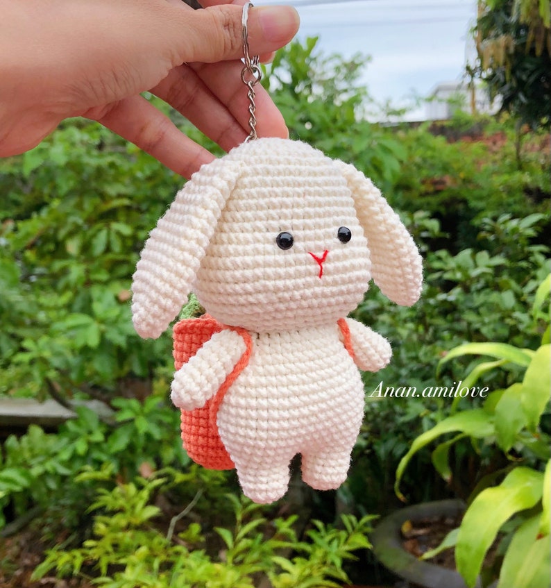 May include: White crocheted bunny keychain with a small orange backpack. The bunny has black eyes and a red nose.