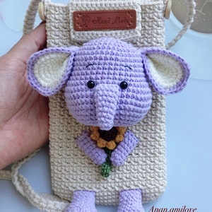 以下が含まれることがあります： 黄色のひまわりを持った紫色の編みぐるみの象が、ベージュの編みぐるみのバッグにストラップで取り付けられています。バッグには、"Hand Made"と書かれた茶色の革のタグが付いています。