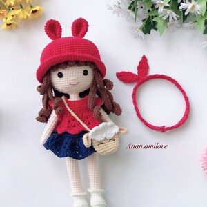 Peut inclure: Une poup&eacute;e en crochet aux cheveux bruns, portant un chapeau rouge avec des oreilles de lapin, un haut rouge et blanc, une jupe bleue et des chaussures blanches. La poup&eacute;e tient un sac en crochet en forme d'agneau. La poup&eacute;e porte &eacute;galement un bandeau rouge.
