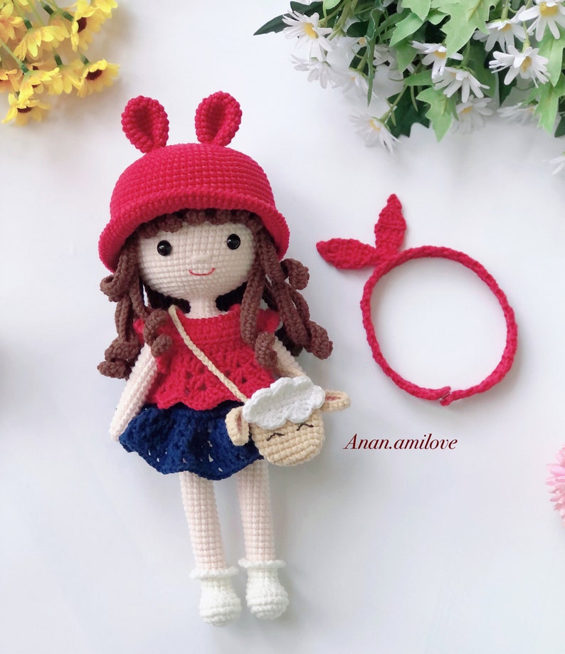 Peut inclure: Une poup&eacute;e en crochet aux cheveux bruns, portant un chapeau rouge avec des oreilles de lapin, un haut rouge et blanc, une jupe bleue et des chaussures blanches. La poup&eacute;e tient un sac en crochet en forme d'agneau. La poup&eacute;e porte &eacute;galement un bandeau rouge.