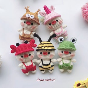 May include: Five crocheted stuffed animals wearing different animal hats and overalls. The animals are a bee, a unicorn, a crab, a squirrel, and a frog. The animals are all white with red, yellow, green, brown, and pink accents.