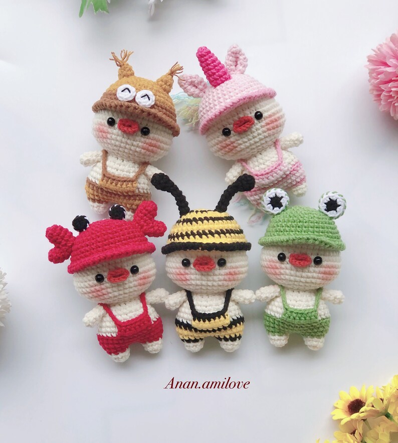 May include: Five crocheted stuffed animals wearing different animal hats and overalls. The animals are a bee, a unicorn, a crab, a squirrel, and a frog. The animals are all white with red, yellow, green, brown, and pink accents.