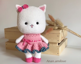 May include: A white crocheted cat wearing a pink dress with a blue trim and a pink bow. The cat is standing on a wooden surface.