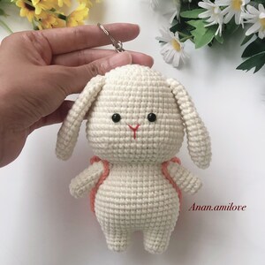 May include: White crocheted bunny keychain with a small orange backpack. The bunny has black eyes and a red nose. The keychain is attached to a silver ring.