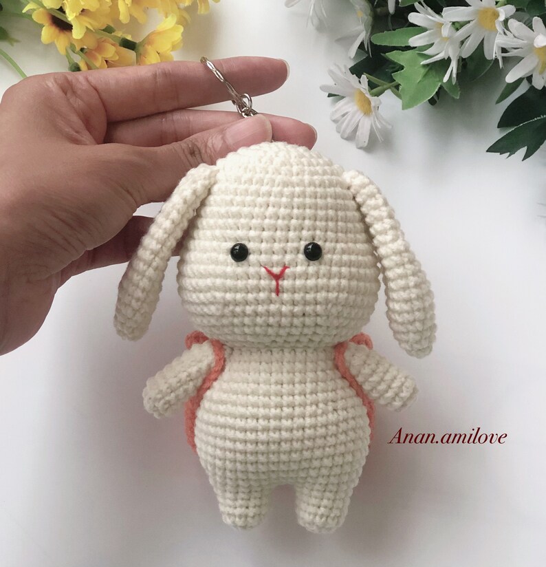 May include: White crocheted bunny keychain with a small orange backpack. The bunny has black eyes and a red nose. The keychain is attached to a silver ring.