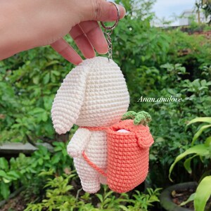 May include: White crocheted bunny keychain with a small orange crocheted bag on its back. The bag has green leaves on top. The keychain is attached to a silver ring.