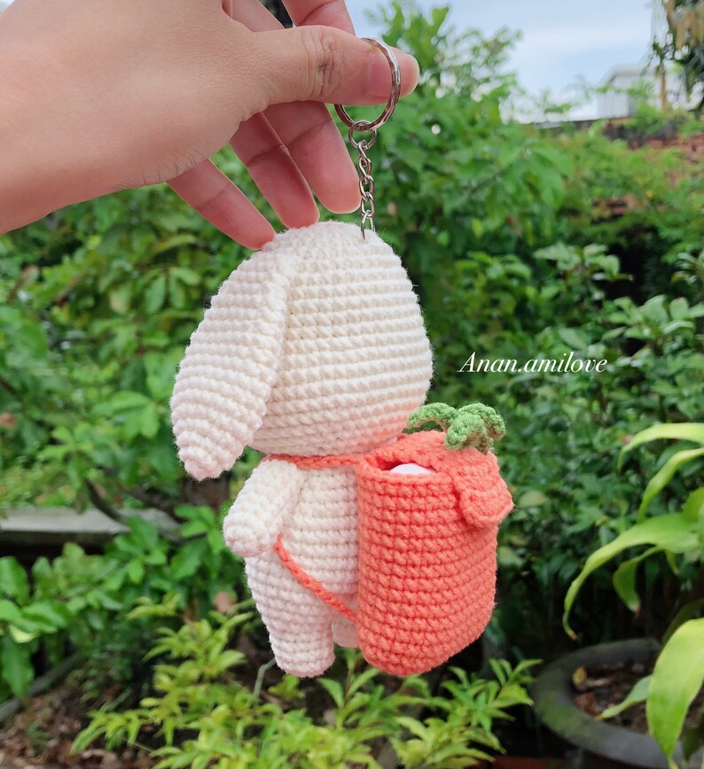 May include: White crocheted bunny keychain with a small orange crocheted bag on its back. The bag has green leaves on top. The keychain is attached to a silver ring.