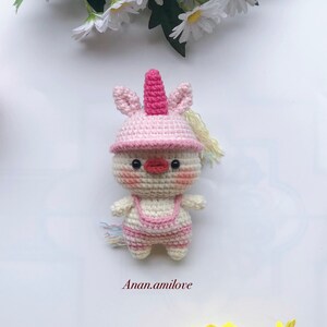 May include: A crocheted unicorn doll with a pink hat and a white body. The doll has a pink horn, pink cheeks, and a red beak. The doll is standing on a white background.