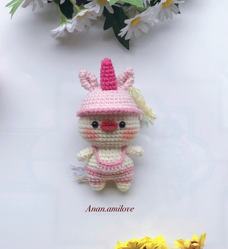 May include: A crocheted unicorn doll with a pink hat and a white body. The doll has a pink horn, pink cheeks, and a red beak. The doll is standing on a white background.