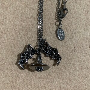 May include: A dark gray necklace featuring a bat wing pendant with a witch hat charm. The bat wings are detailed with small black accents. The necklace has a delicate chain and a lobster clasp.