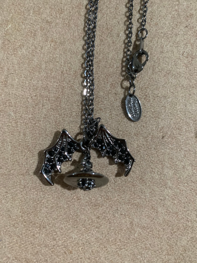 May include: A dark gray necklace featuring a bat wing pendant with a witch hat charm. The bat wings are detailed with small black accents. The necklace has a delicate chain and a lobster clasp.