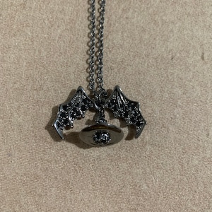 May include: A silver-toned necklace with a bat wing pendant and a circular pendant with black stones. The bat wings are detailed with black stones. The necklace is displayed on a neutral-toned surface.