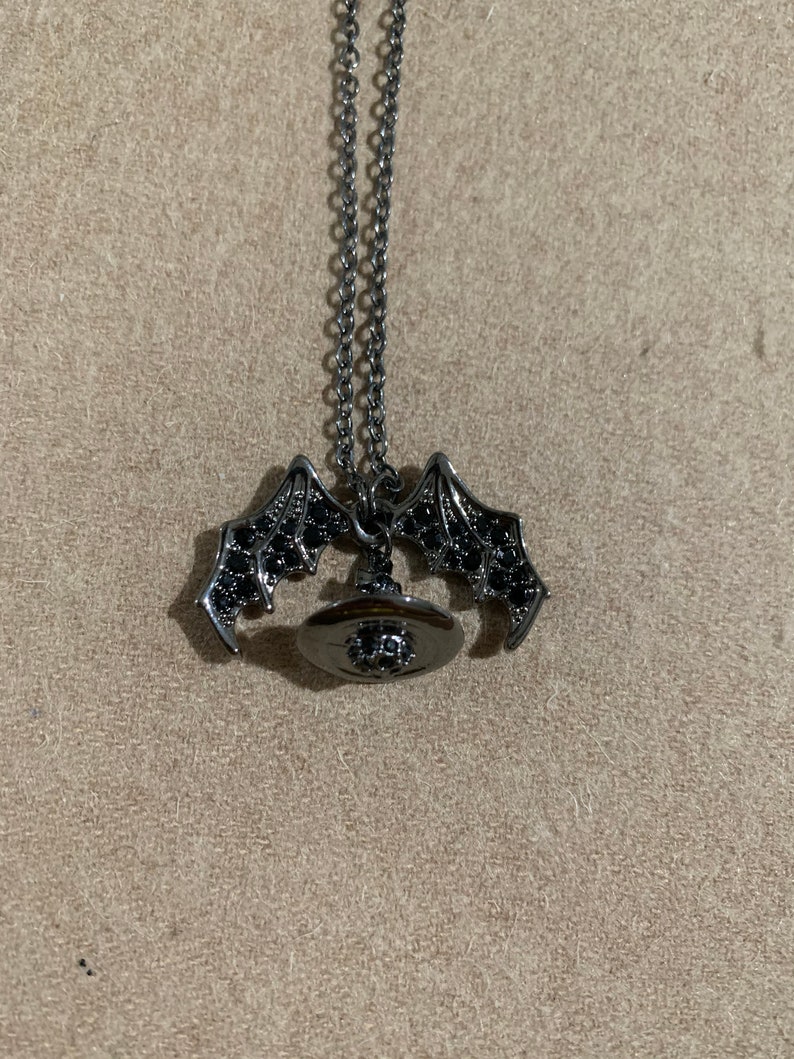 May include: A silver-toned necklace with a bat wing pendant and a circular pendant with black stones. The bat wings are detailed with black stones. The necklace is displayed on a neutral-toned surface.
