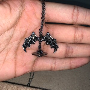 May include: A dark gray necklace featuring a bat wing pendant. The pendant has a detailed design with black accents. The chain is a matching dark gray color. The necklace is displayed against a neutral background.