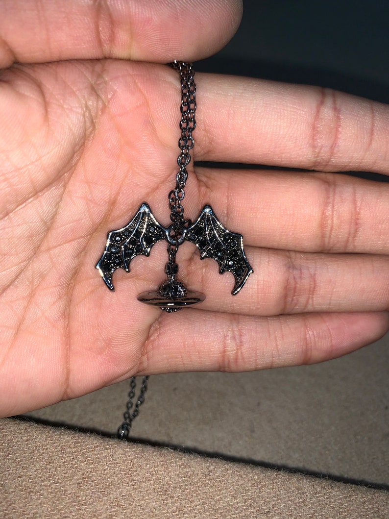 May include: A dark gray necklace featuring a bat wing pendant. The pendant has a detailed design with black accents. The chain is a matching dark gray color. The necklace is displayed against a neutral background.