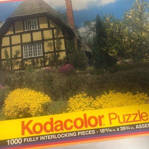 May include: A 1000 piece jigsaw puzzle featuring a quaint, white and brown, half-timbered cottage with a thatched roof. The cottage is surrounded by lush green foliage and colourful flowers. The puzzle box has the text "Kodacolor Puzzle" and "1000 FULLY INTERLOCKING PIECES - 18 15/16 in. X 26 3/4 in. ASSEMBLED".