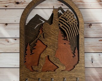 May include: Wooden key holder with a cutout design of a Bigfoot holding an American flag in front of a mountain range. The key holder has five hooks.