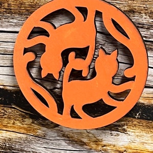 May include: Orange wooden ornament with a cutout design of two cats. The ornament is hanging from a black cord.