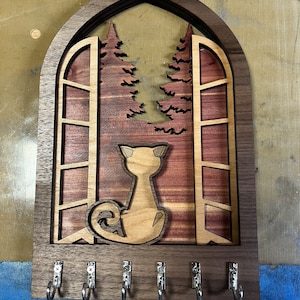 May include: A wooden key holder shaped like a window with a cat silhouette inside. The window has a wood grain pattern and is surrounded by a dark wood frame. There are five metal hooks on the bottom for hanging keys.