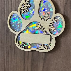 May include: A wooden paw print ornament with a holographic finish. The ornament has a blank space for personalization and features snowflakes cut out around the edges.