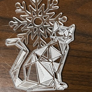 May include: A clear acrylic Christmas ornament featuring a geometric cat design with a snowflake behind it.