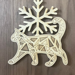 May include: A wooden Christmas ornament featuring a geometric cat design with a snowflake above it. The ornament is made of light brown wood and has a red string for hanging.