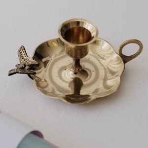 Könnte beinhalten: Ein goldfarbener Kerzenhalter mit Schmetterlingsmotiv. Der Halter hat einen gewellten Rand und einen kleinen Griff.