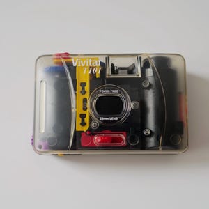 May include: A clear plastic Vivitar T101 disposable camera. The camera has a black body with yellow and red accents. The lens is labelled "28mm LENS" and "FOCUS FREE". The camera is designed for single-use.
