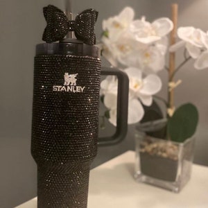 Puede incluir: Un vaso de acero inoxidable negro con un acabado de brillo negro y un lazo negro en la tapa. El vaso tiene un asa y el logotipo de Stanley es visible en el lateral.