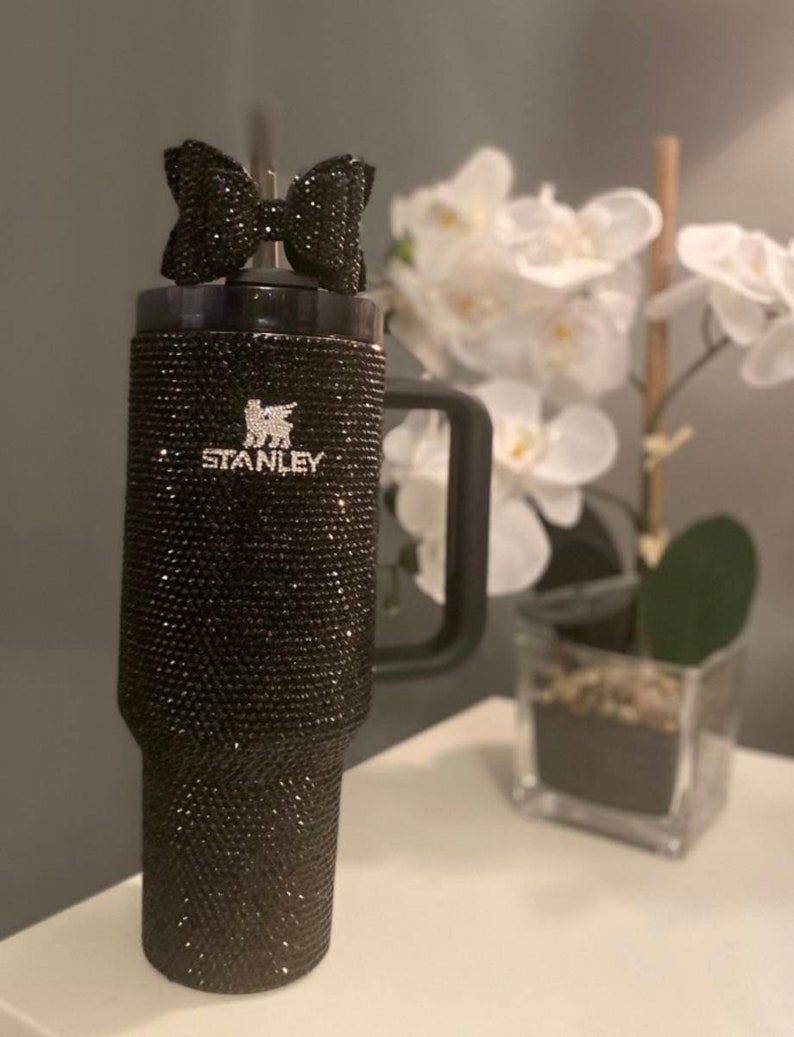 Puede incluir: Un vaso de acero inoxidable negro con un acabado de brillo negro y un lazo negro en la tapa. El vaso tiene un asa y el logotipo de Stanley es visible en el lateral.