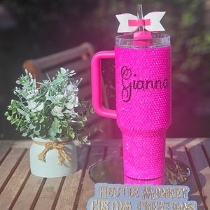 May include: Pink glitter tumbler with a handle and a white bow on the lid. The tumbler is personalized with the name "Gianna". The tumbler is on a wooden surface with a blue sign that says "Hustle Monkey Custom Creations".
