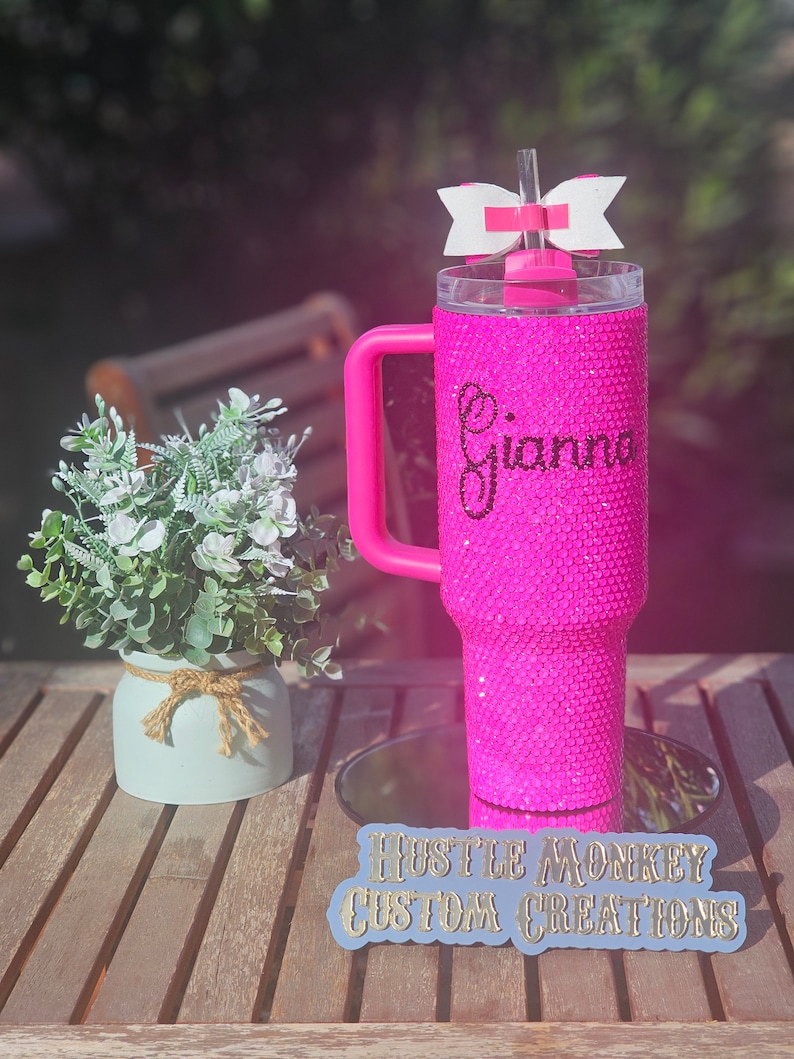 May include: Pink glitter tumbler with a handle and a white bow on the lid. The tumbler is personalized with the name "Gianna". The tumbler is on a wooden surface with a blue sign that says "Hustle Monkey Custom Creations".